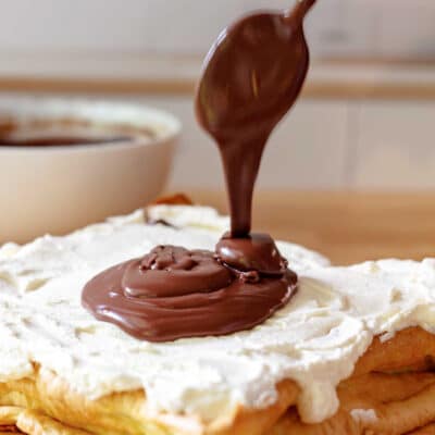 A square eclair cream puff cake on butcher block and spread with whipped cream. A spoon is drizzling a pool of ganache on top.