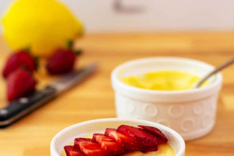 Two white ramekins of lemon pudding, one with thinly sliced red strawberries on top. There are strawberries and a lemon in the background.