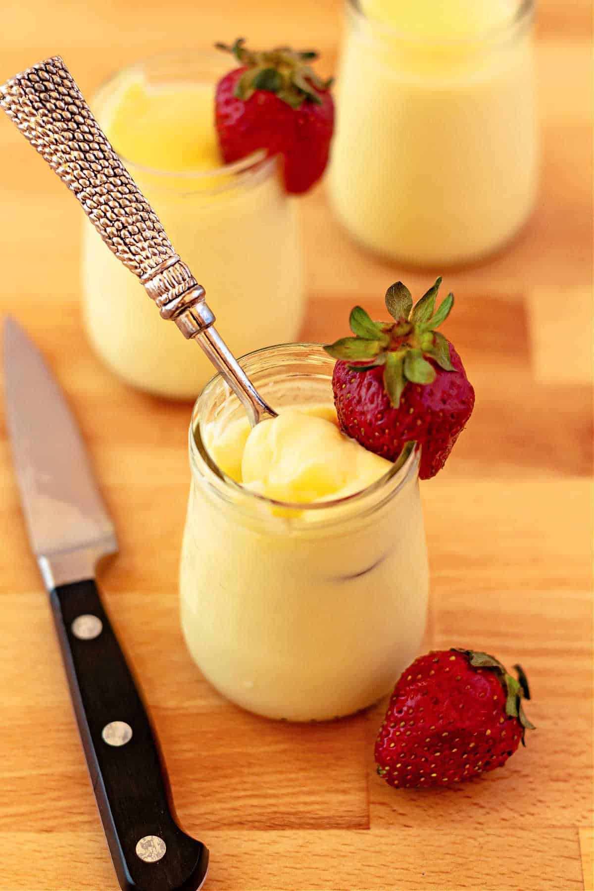 Three small clear glass jars filled with pale yellow lemon pudding. Two of the jars have a slit whole strawberry on their rims.