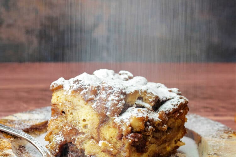 A square slice of bread pudding on a marbled plate with powdered sugar sifting down on top of it.