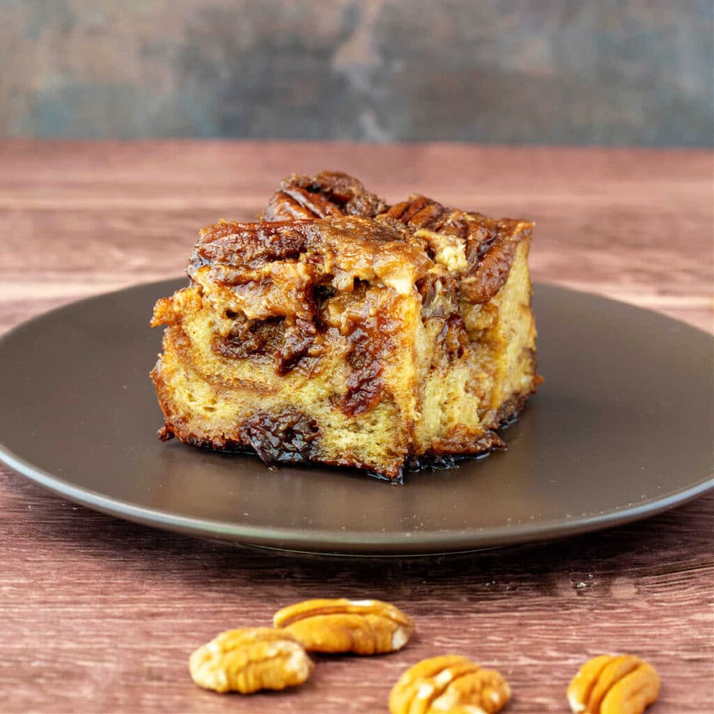 A square of bread pudding on a brown plate with pecans in the foreground.