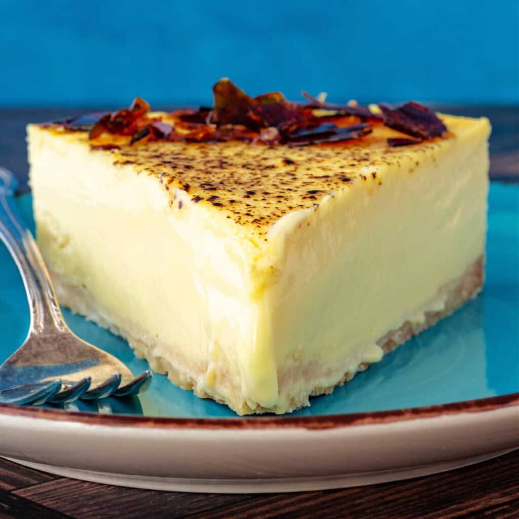 A large slice of pale yellow cheesecake decorated with shards of caramelized sugar on a blue plate.