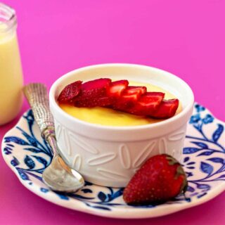 A square image of a white ramekin of lemon pudding and sliced strawberries on a blue floral plate shot on a bright pink background.