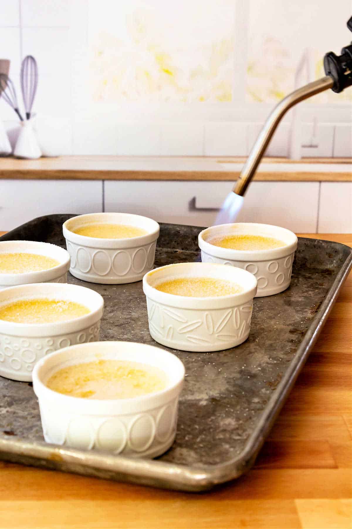 A tray of creme brulee in white ramekins. The nozzle of a blowtorch can be seen in the upper right with a blue flame coming out, melting and caramelizing the sugar on one of the desserts.
