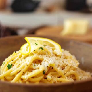 A mound of lemon garlic pasta topped with grated cheese and a thin slice of lemon in a light brown bowl.