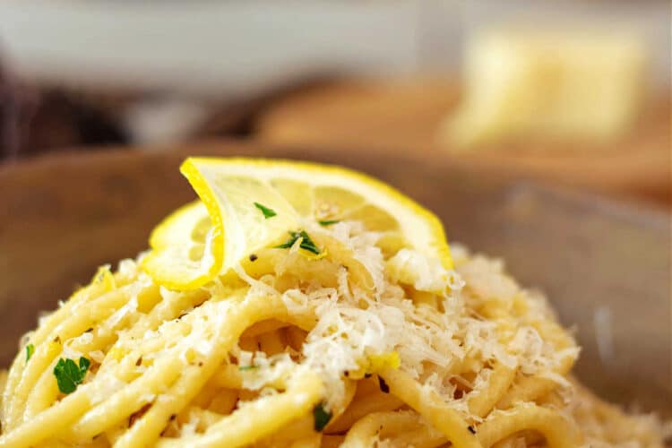 A mound of lemon garlic pasta topped with grated cheese and a thin slice of lemon in a light brown bowl.