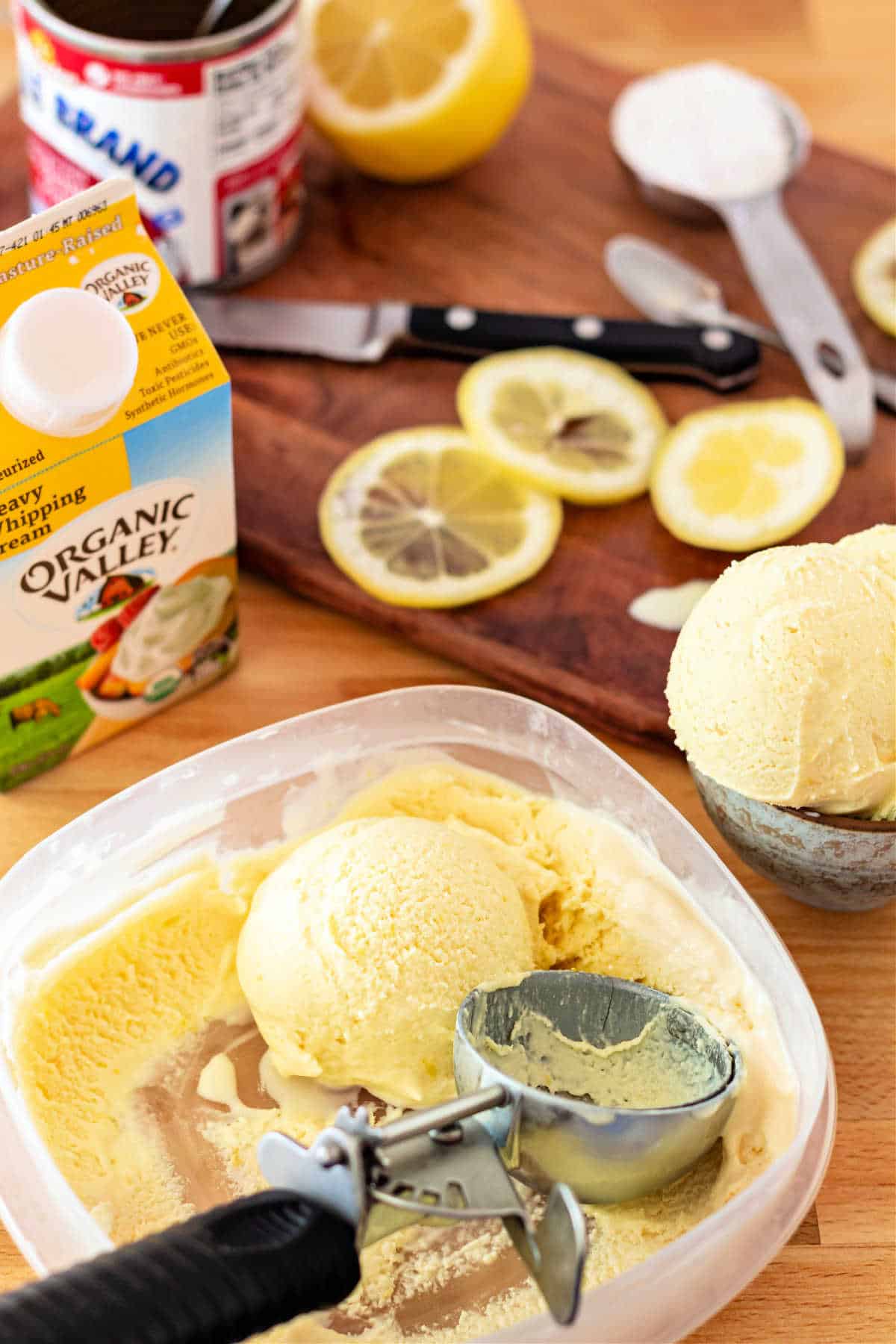 A container of ice cream with an ice cream scoop in it. ON the counter are a carton of heavy cream, lemon slices, a spoonful of powdered sugar, and a can of sweetened condensed milk.