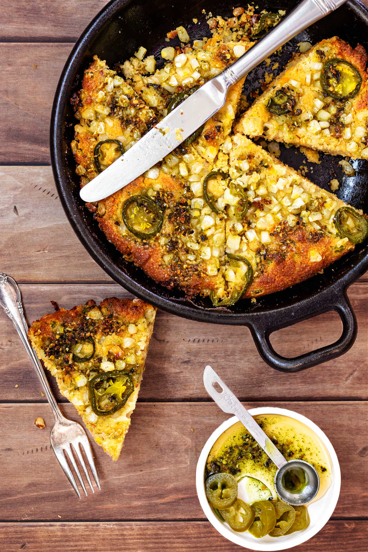 An overhead shot of a skillet of cut-up cornbread with jalapenos on top, a slice of the cornbread on a wooden surface and a white dish with chili crisp, hot honey, and pickled jalapenos in it.
