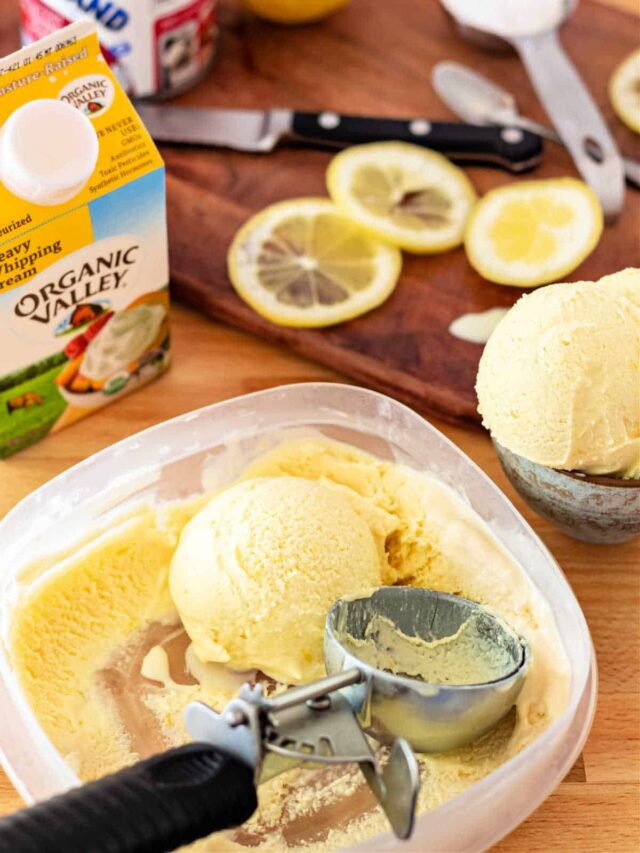 A container of ice cream with an ice cream scoop in it. ON the counter are a carton of heavy cream, lemon slices, a spoonful of powdered sugar, and a can of sweetened condensed milk.