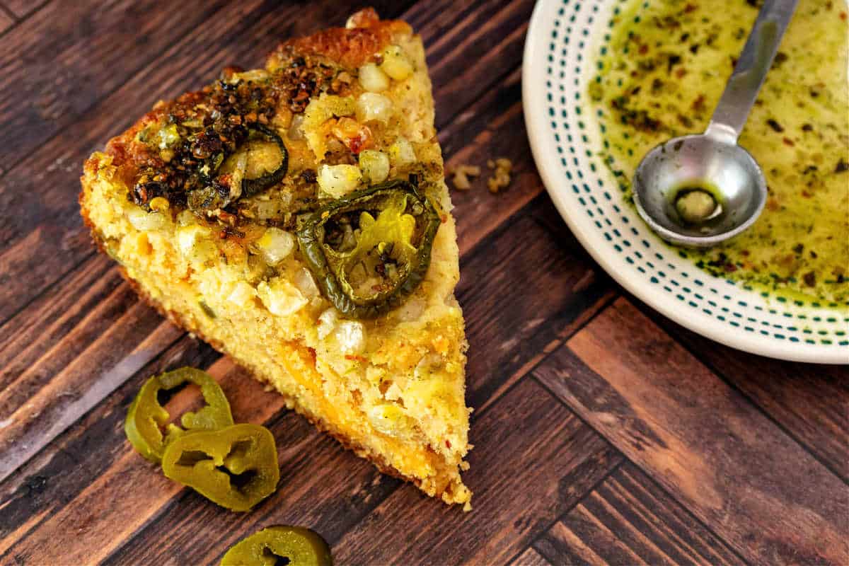 A wedge of cornbread topped with rings of jalapeno with a dish of honey and green chili crisp next to it.