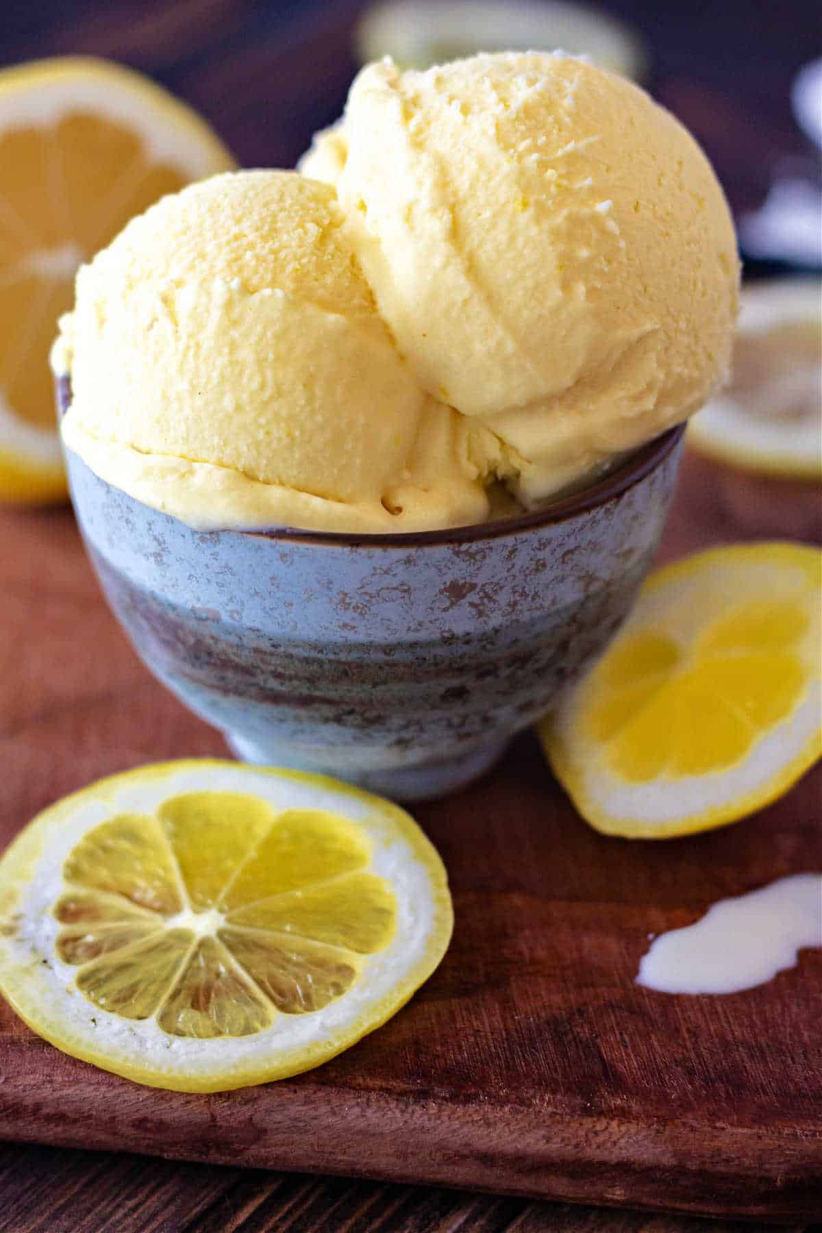 A small blue bowl with scoops of lemon ice cream in it and lemon slices on a wooden surface.