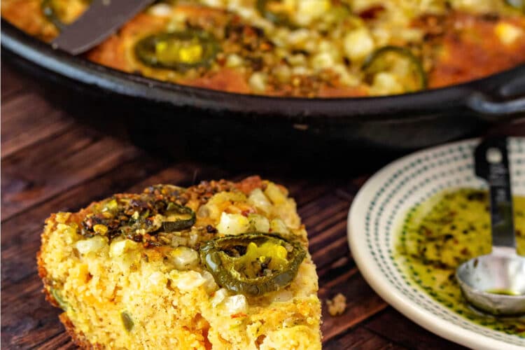 A slice of loaded cornbread with pickled jalapenos on top and next to it on a wooden surface with the skillet containing the rest of the cornbread in the background.