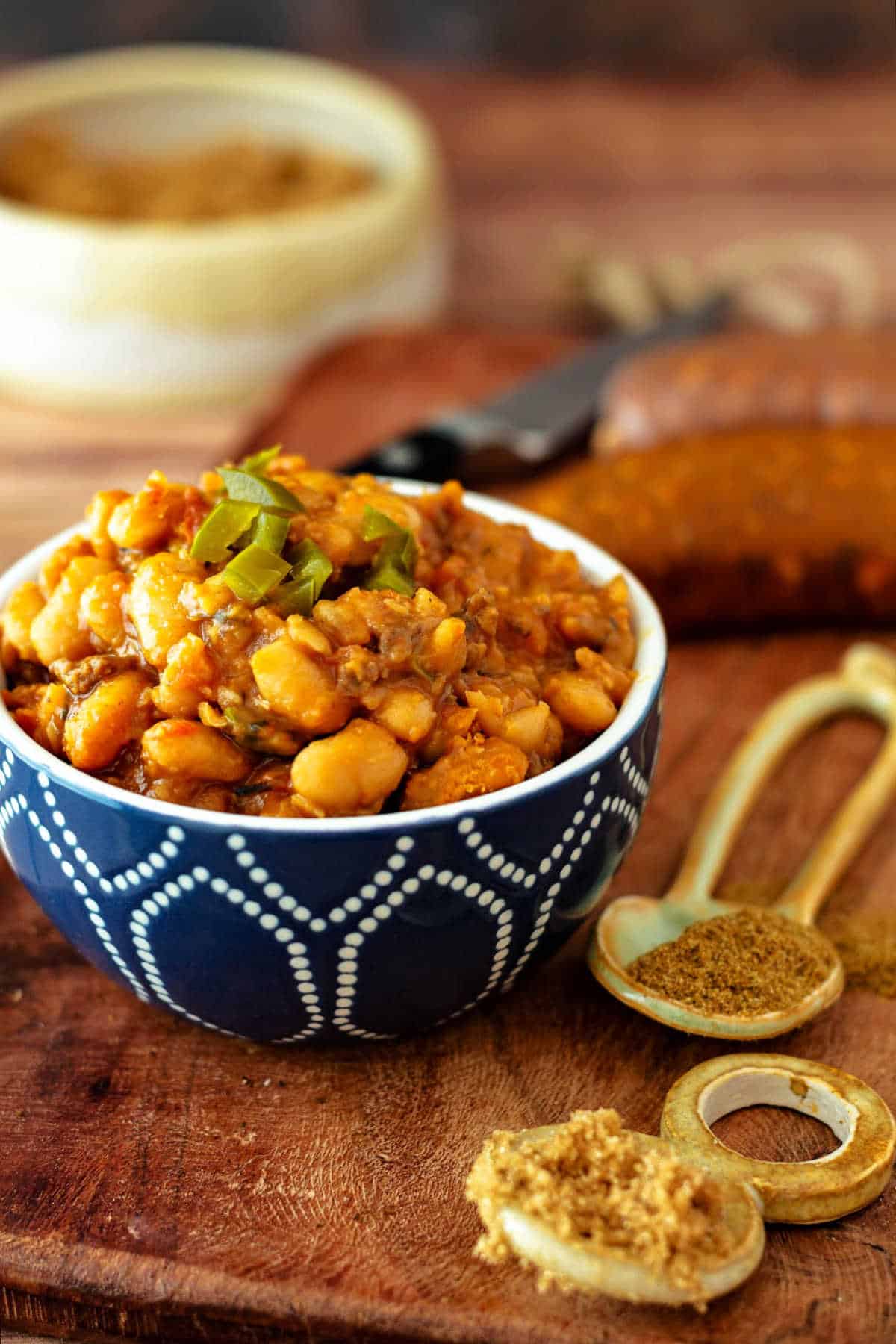 A small blue bowl of baked beans with spoonfuls of brown sugar and cumin next to it with some uncooked chorizo in the background.