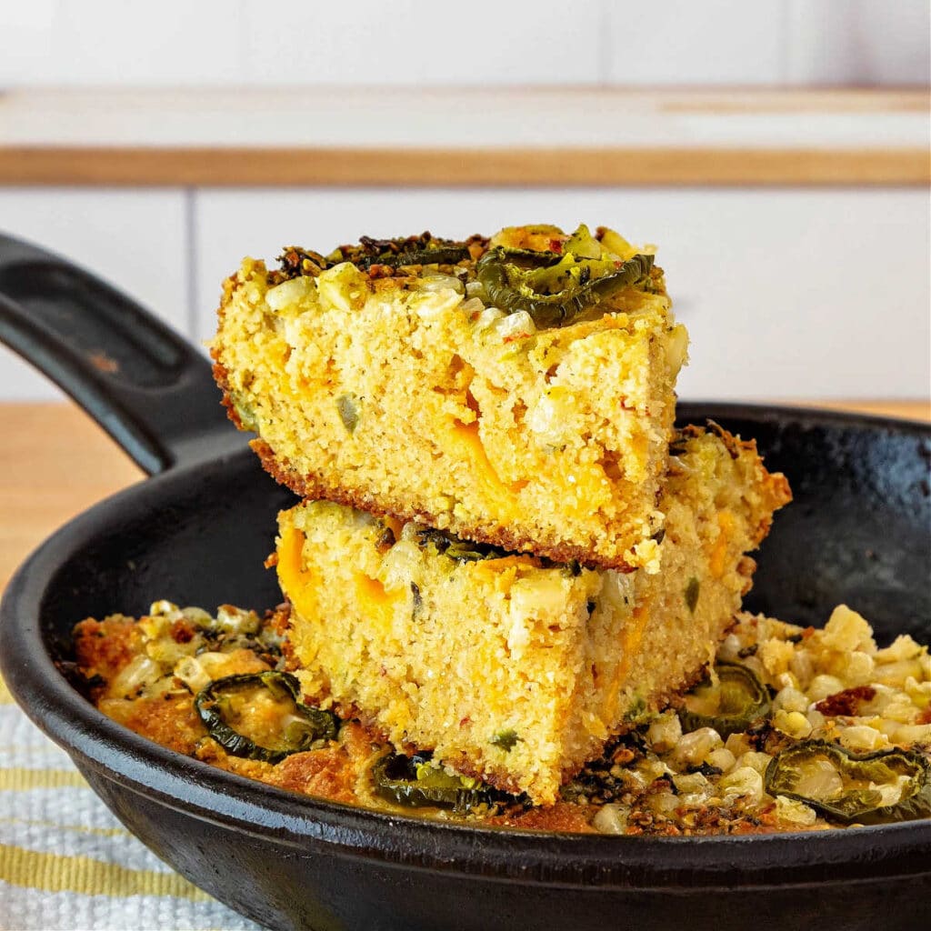 A skillet of cornbread topped with jalapeno peppers. Two sliced are cut and stacked on top of the rest of the cornbread.