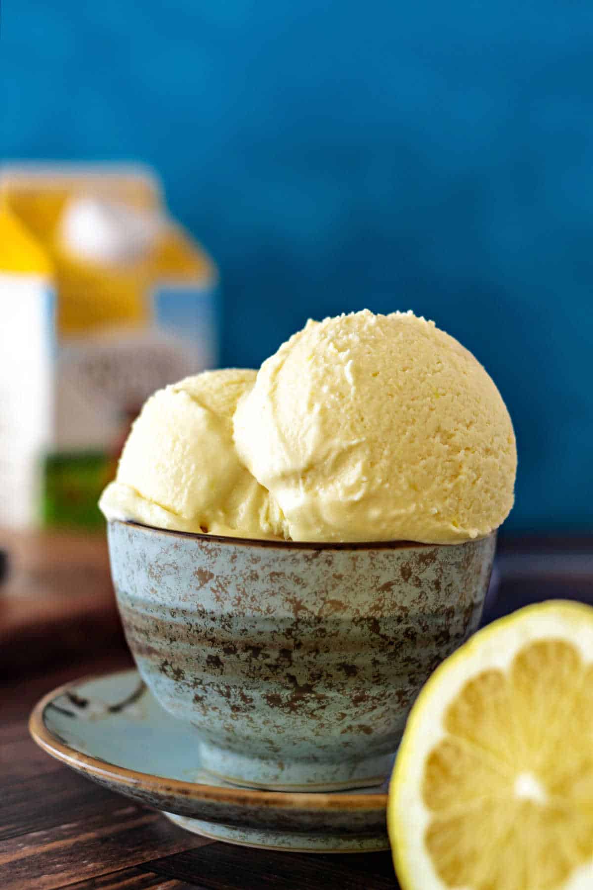 Two scoops of lemon ice cream in a pale blue bowl with a carton of heavy cream in the background and a cut lemon in the foreground.