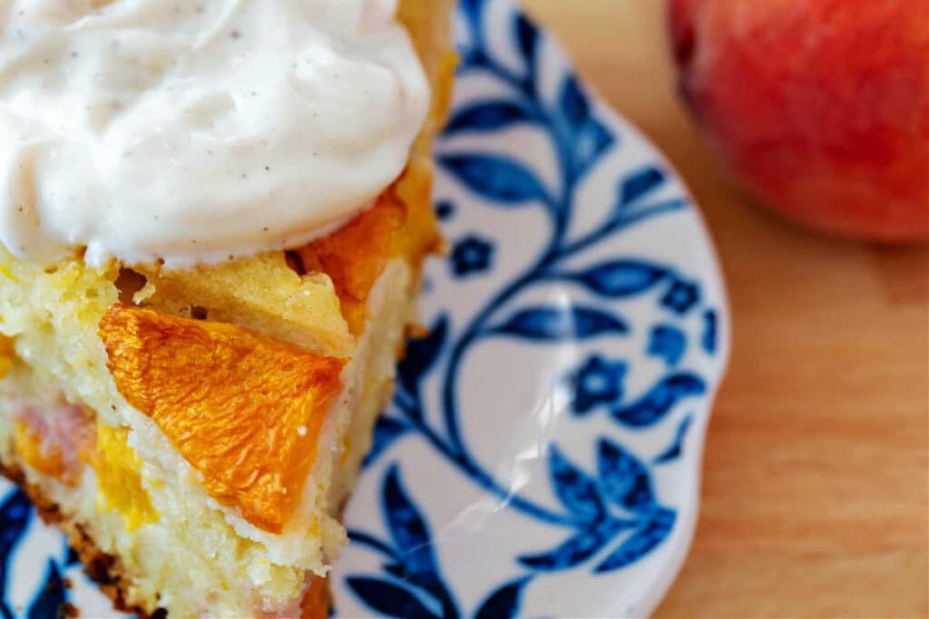 A close-up, overhead view of a slice of peach cake on a blue and white patterned plate with a dollop of vanilla yogurt on top.