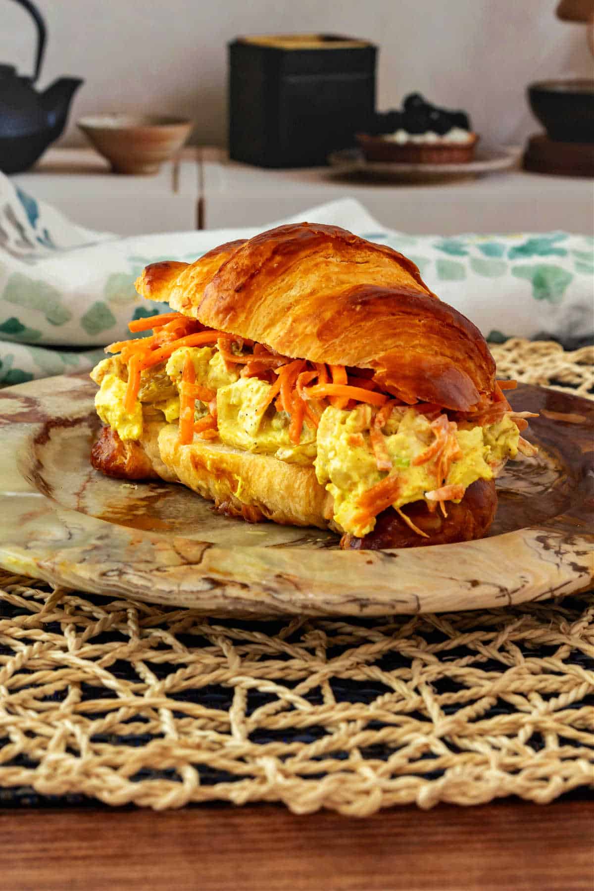A chicken salad sandwich with shredded carrots made on a split croissant and on an earth-toned plate.
