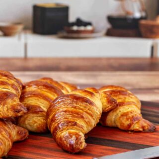 Several shiny and deep golden brown croissasnts on a cutting board with a bread knife.