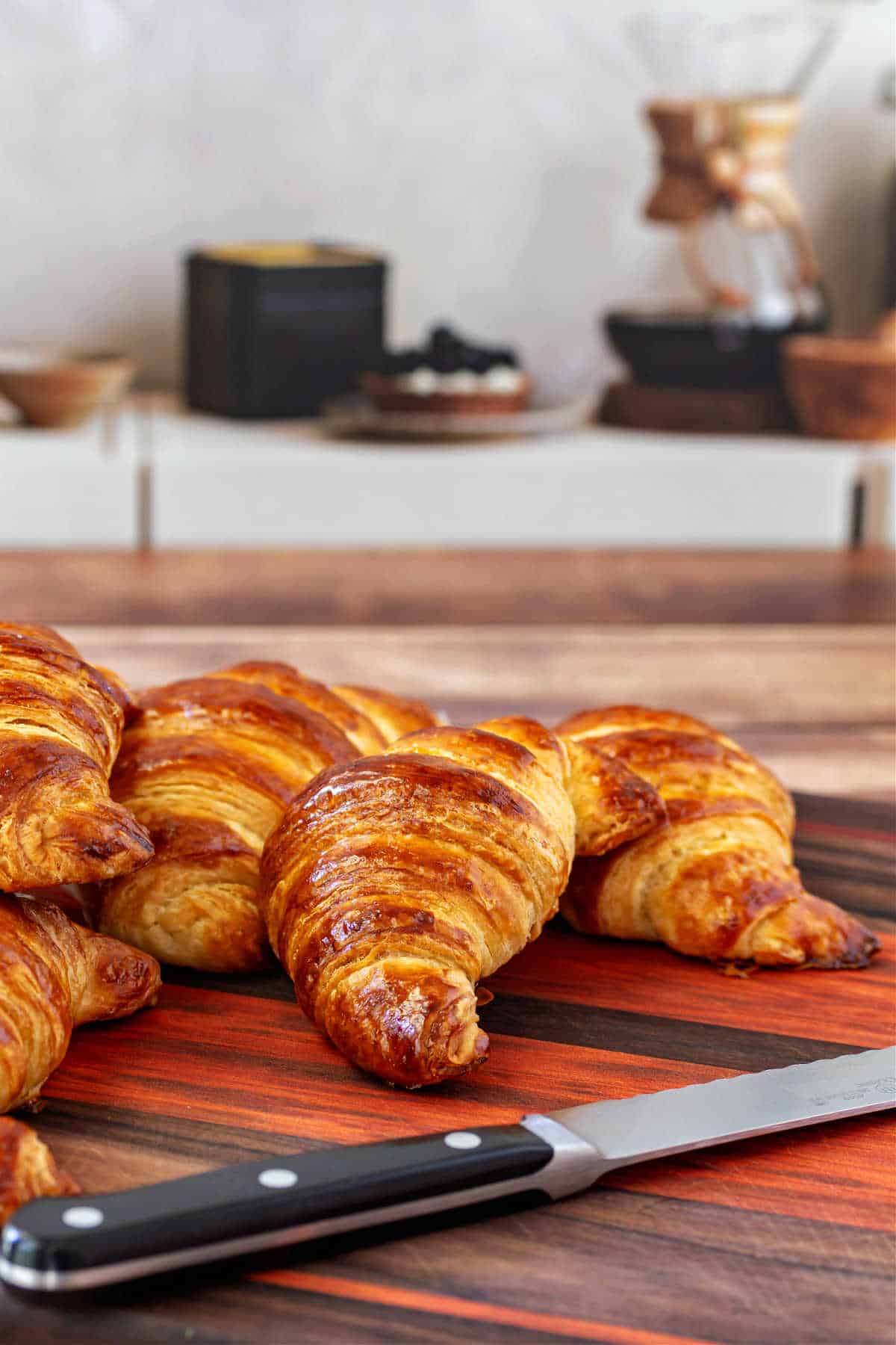 Several shiny and deep golden brown croissasnts on a cutting board with a bread knife.