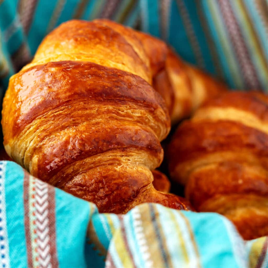 A close up of two deep, golden-brown croissants in a basket lined with a light blue and white striped linen cloth.