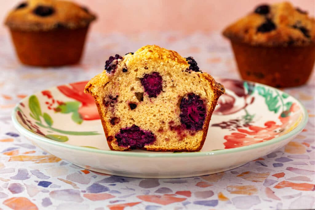 A cross-section of a jumbo blackberry muffin on a plate showing lots of blackberries in the muffin.