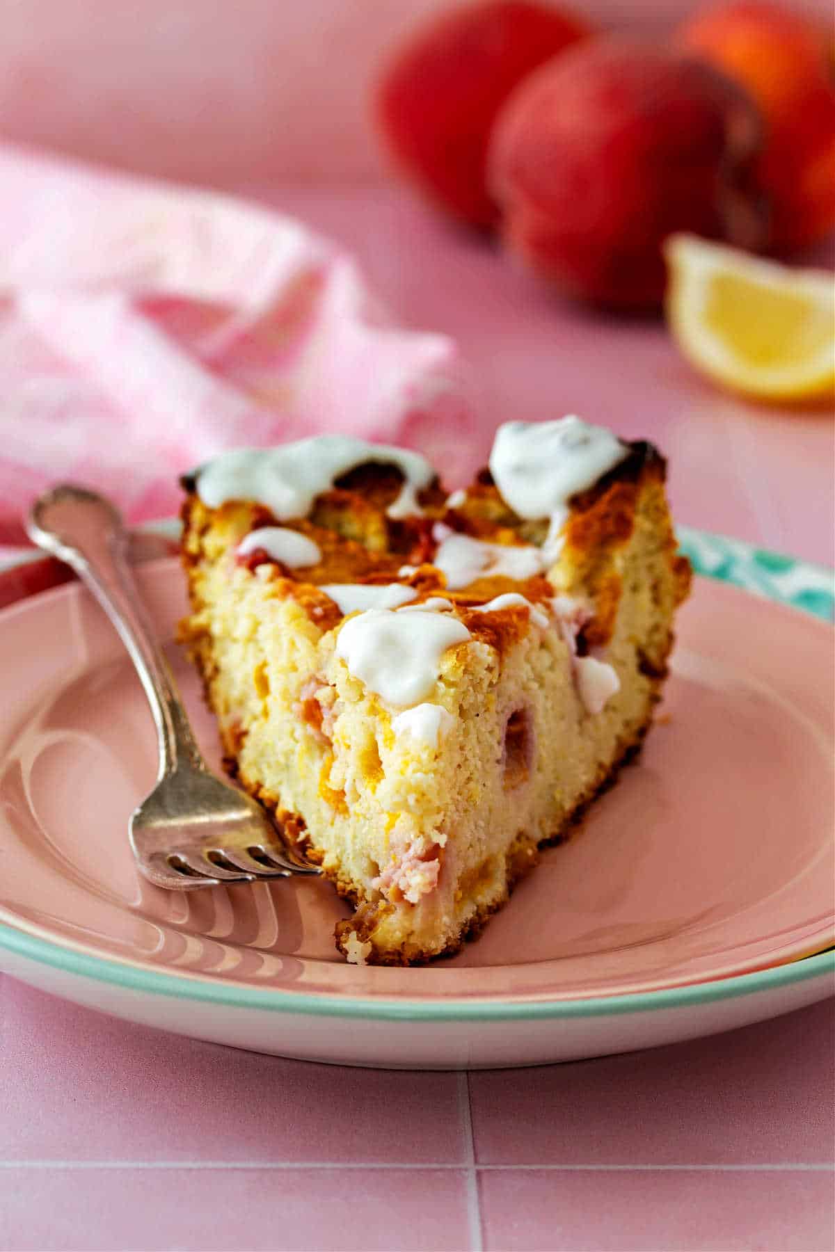 A slice of cake with lots of pieces of peaches in it and on top on a pink plate with 3 peaches and a slice of lemon in the background.