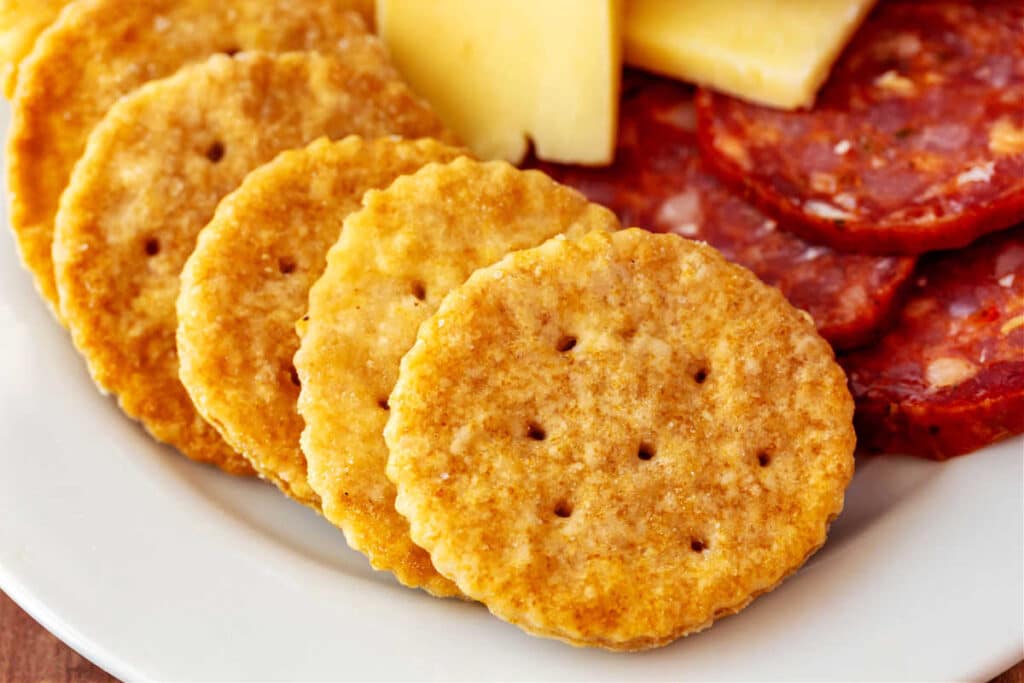 A close up of about half of a white plate with some homemade Ritz crackers, slices of salami, and pieces of pale yellow cheese on it.