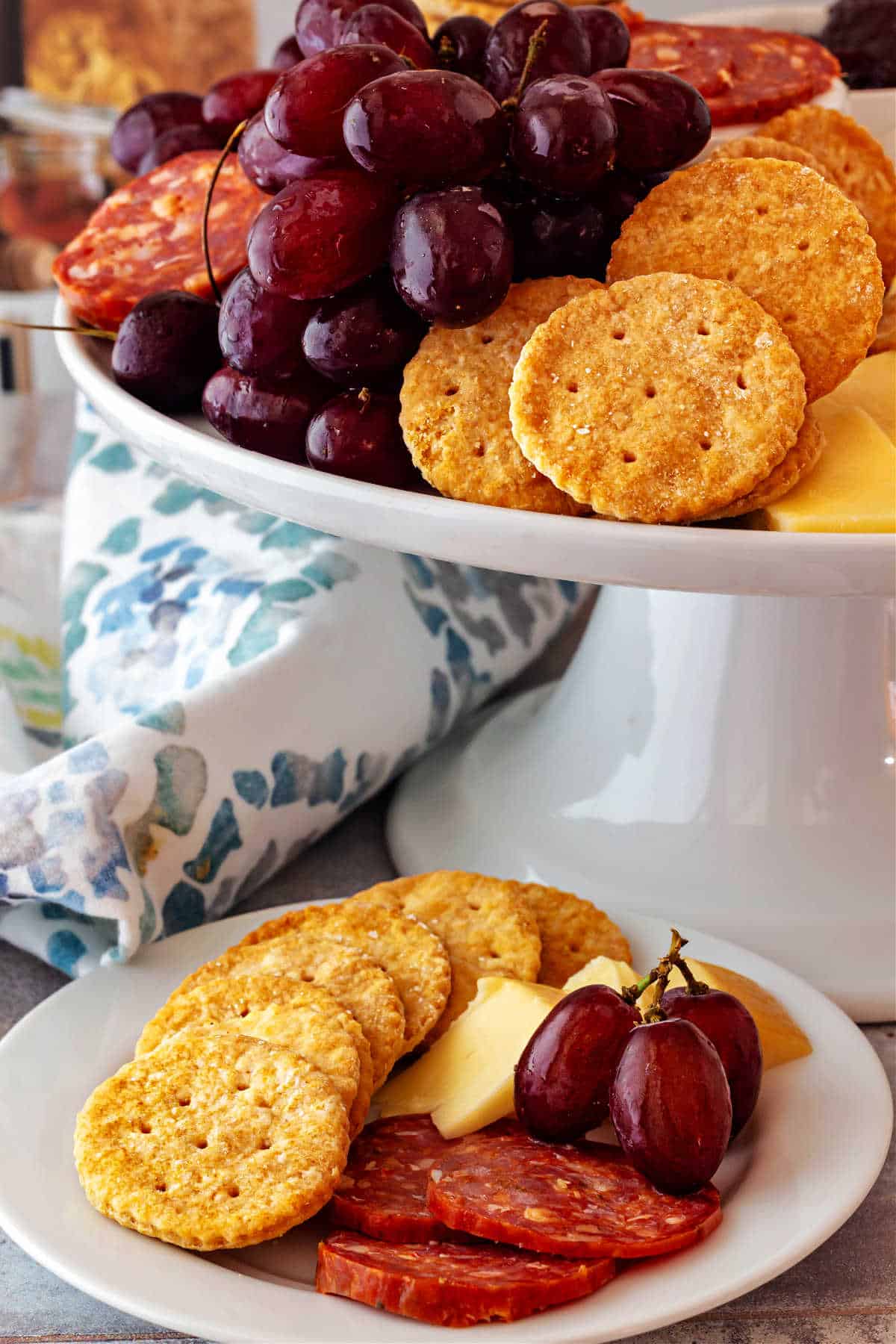 A white, ceramic cake stand folled with red grapes, cheese, and Ritz crackers. There is a small white plate next to it with Ritz, sliced sausage, some cheese, and 3 red grapes.