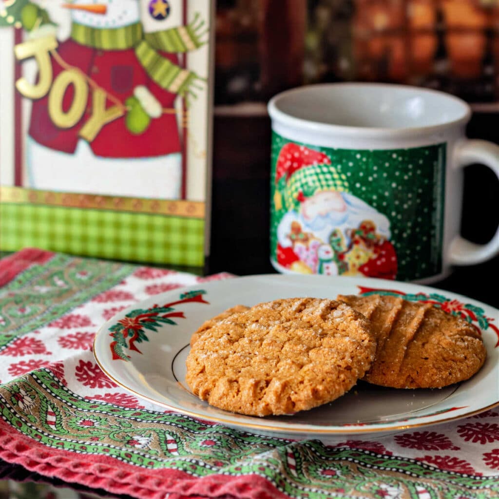 Several peanut butter cookies on a white plate with red and green ribbons around the border. There is a Santa mug in the background as well as a greeting card with a snowman that reads, "Joy." 