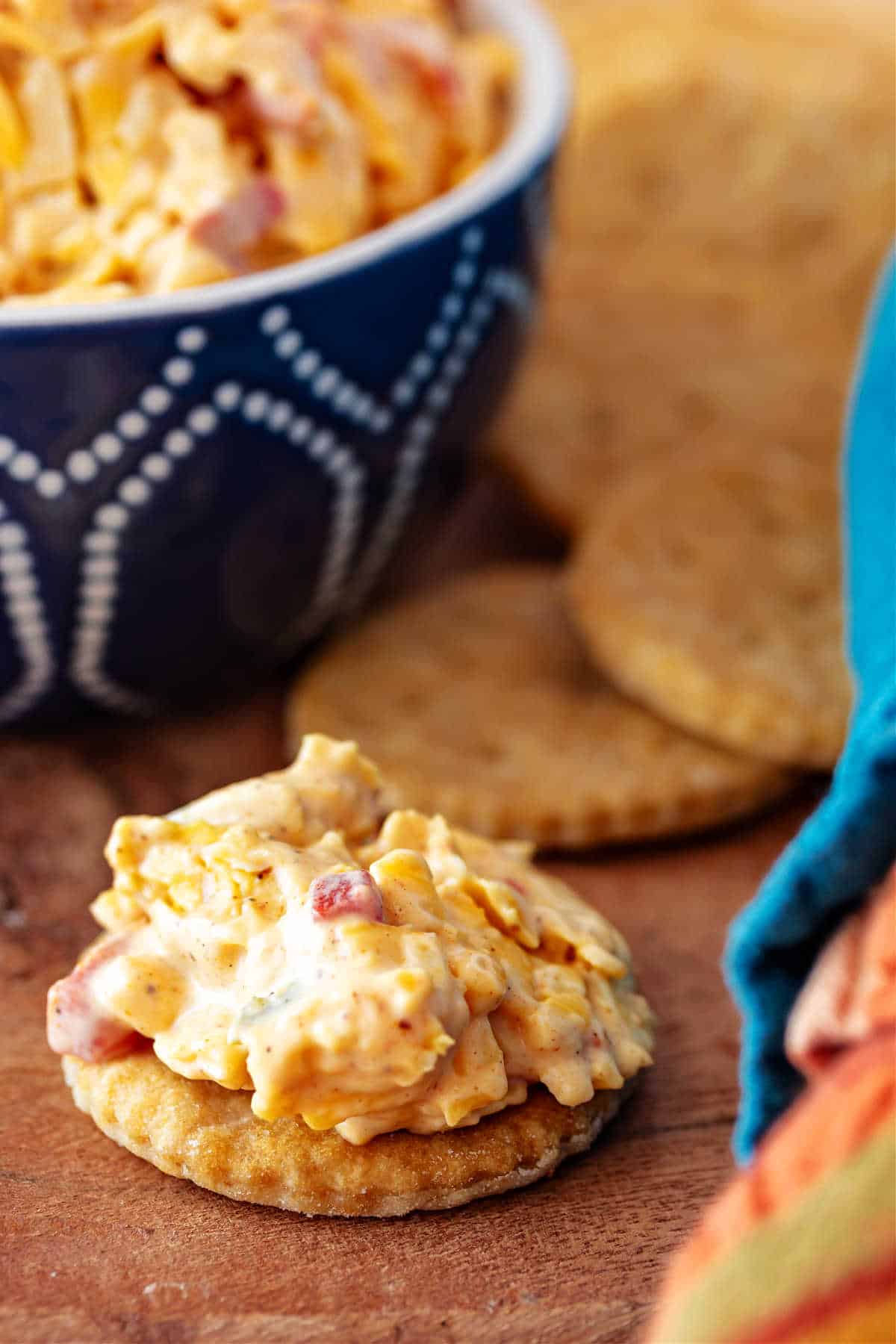 A cracker with a dollop of pimento cheese on top. There is also a blue bowl with white dots to the back left, partially out of the frame, filled with more pimento cheese.