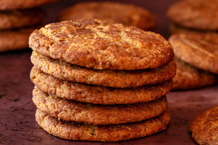 A stack of snickerdoodle cookies with a ceramic spoon of ground cinnamon in the foreground. There are more cookies cooling on a rack in the background.