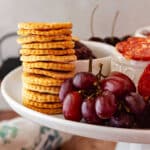 A tall stack of homemade Ritz crackers on a white, rimmed platter next to a bunch of red grapes and sliced cheese.