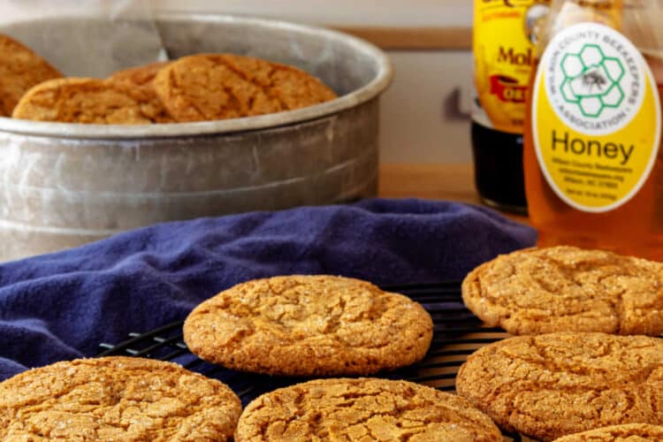 Ginger cookies cooling on a round, wire rack with an aluminum container or more cookies in the background along with a jar of honey and one of molasses.
