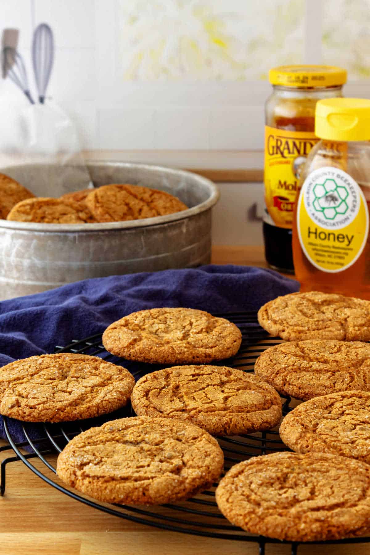 Ginger cookies cooling on a round, wire rack with an aluminum container or more cookies in the background along with a jar of honey and one of molasses.