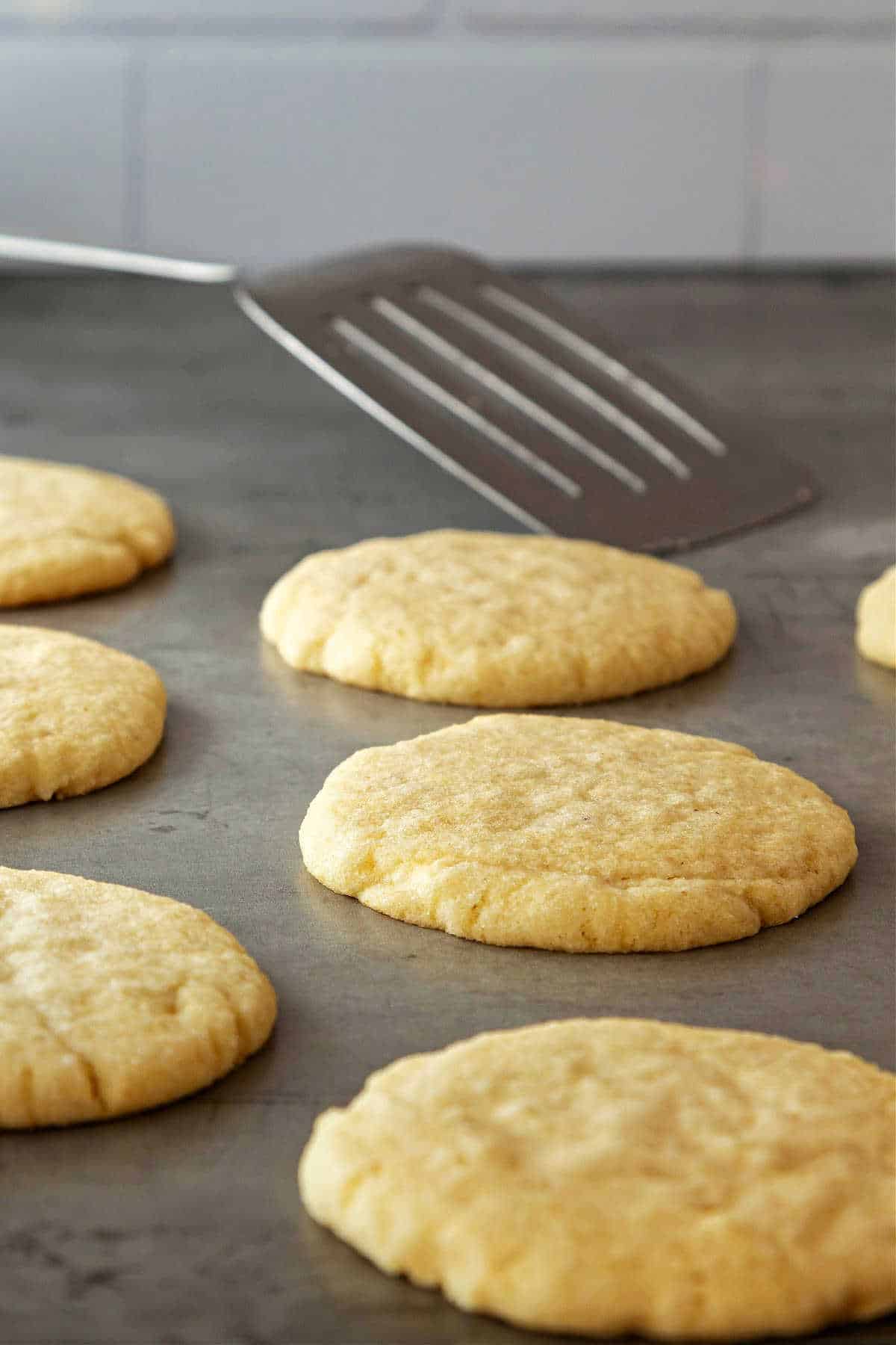 Several large, round sugar cookies on a sheetpan with a metal turner/spatula in the background.
