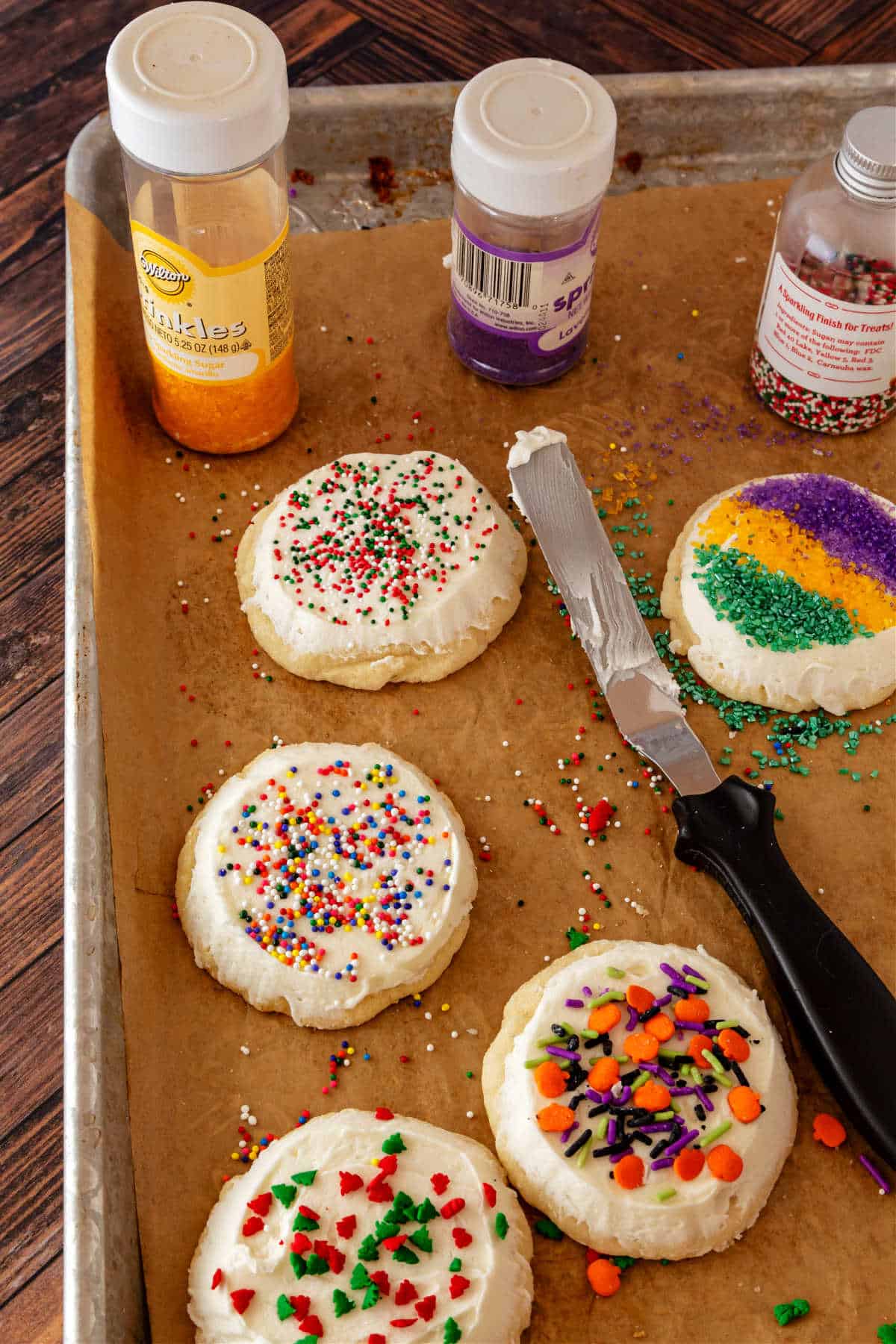 A sheetpan with a piece of beige parchment lining it. On the sheet are several cookies frosted with white icing and decorated with different colors of sprinkles for different holidays. There is also a small icing spatula on the tray with 3 bottles of sprinkles.