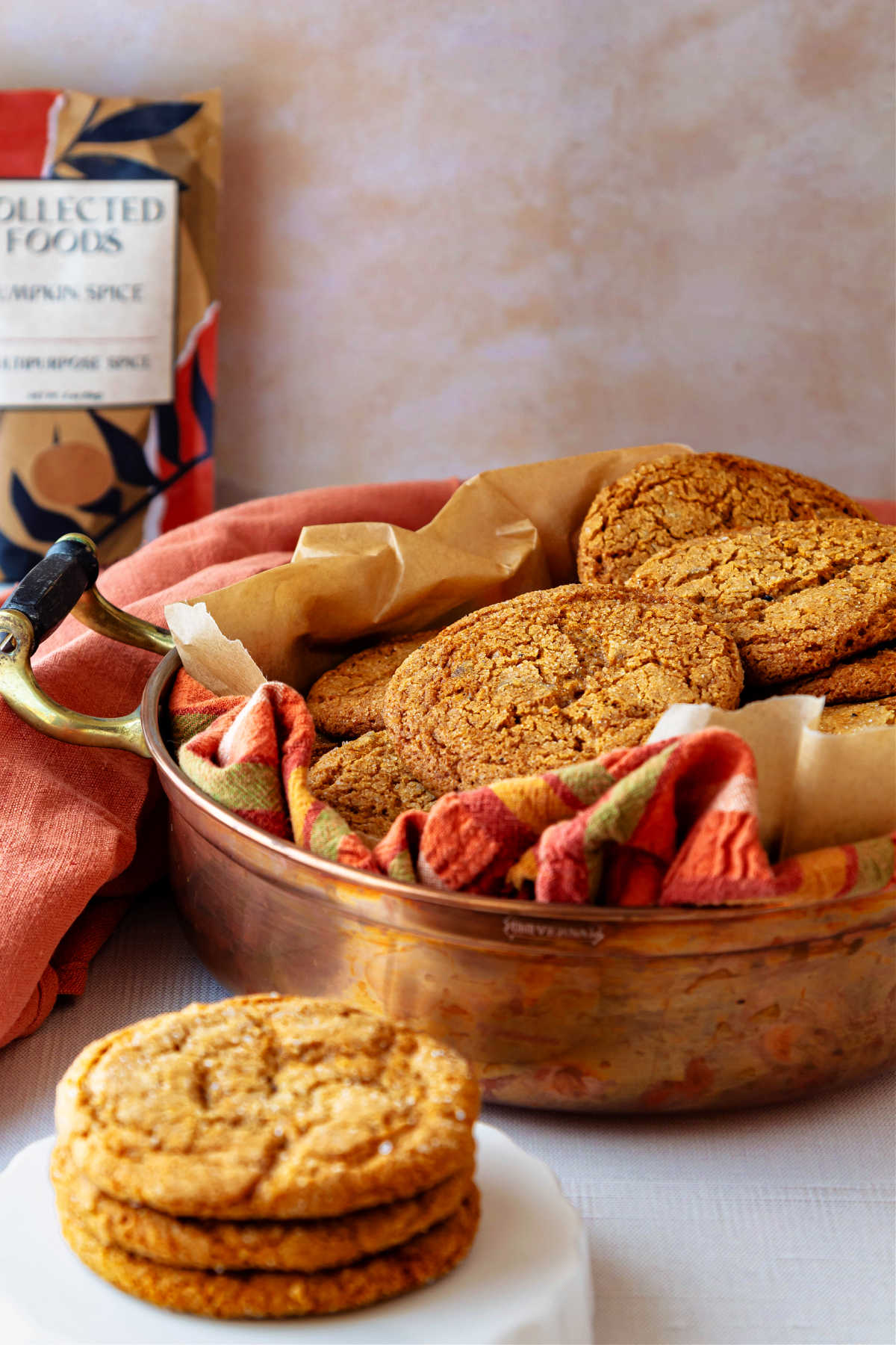 A copper pan lined with an orange-striped napkin with crackled-topped chewy ginger cookies in it. There are 3 cookies stacked in front of the pan on a white riser and a packet of pumpkin spice in the background.