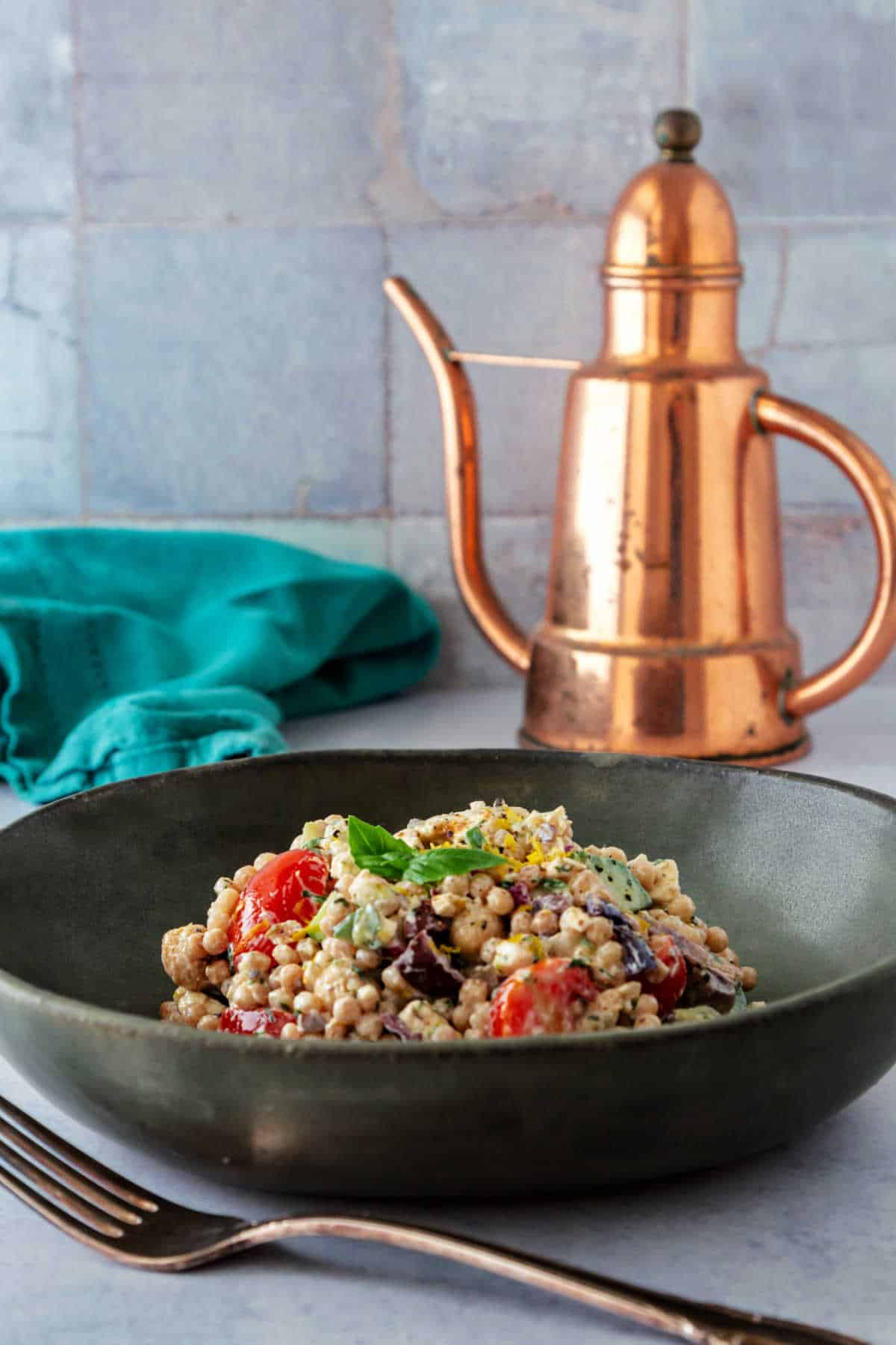 A dark gray bowl of pearl couscous salad on a gray surface with a gray tile backdrop and a copper oil cruet in the background.