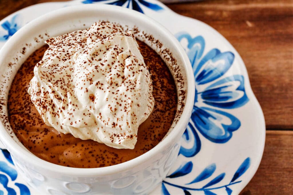 A white ramekin of chocolate mousse topped with coffee whipped cream on a blue-and-white flowered plate. The whipped cream is dusted with cocoa powder.