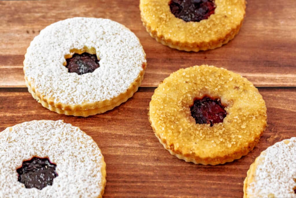 Round Linzer cookies on a wooden surface. All have deep red jam in the centers. Two of them are topped with powdered sugar and the other two with sparkly sugar in the raw.
