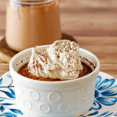 A ramekin of chocolate mousse topped with whipped cream with another glass jar of mousse with a spoon in it in the background.