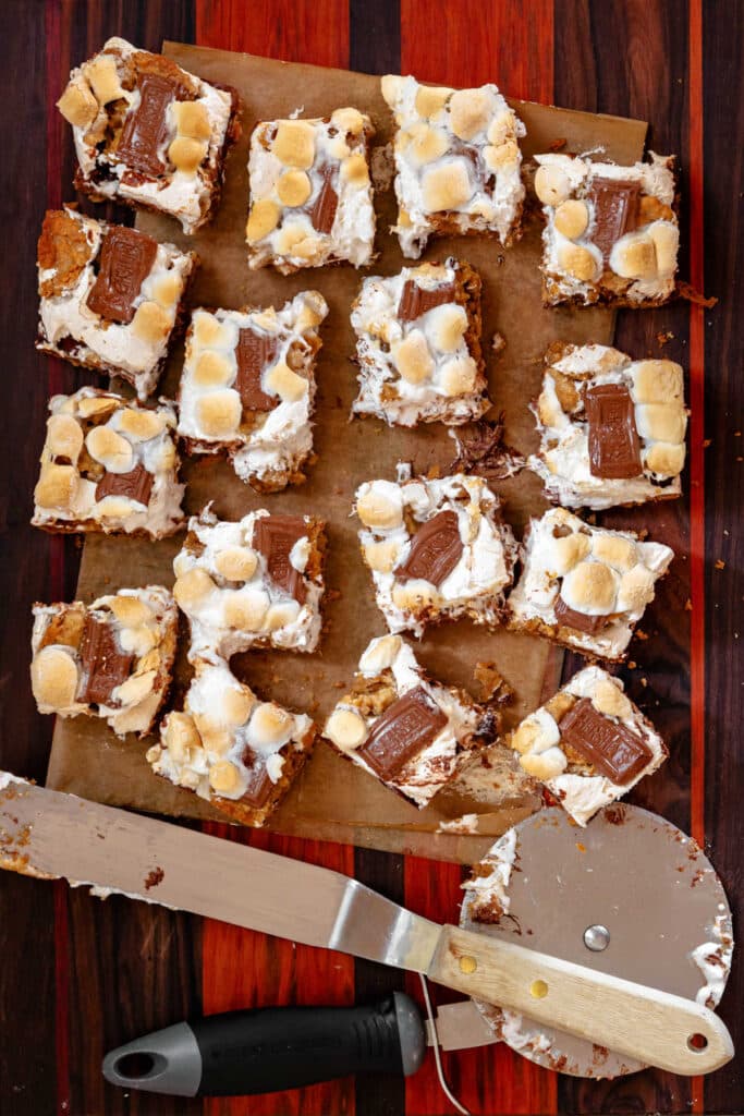 An overhead shot of a large slab of s'mores bar cookies cut into 16 pieces on a piece of parchment with an offset spatula and a pizza wheel at the bottom.
