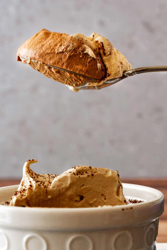 A spoonful of chocolate mousse held over a white ramekin with the rest of the whipped cream-topped chocolate mousse.