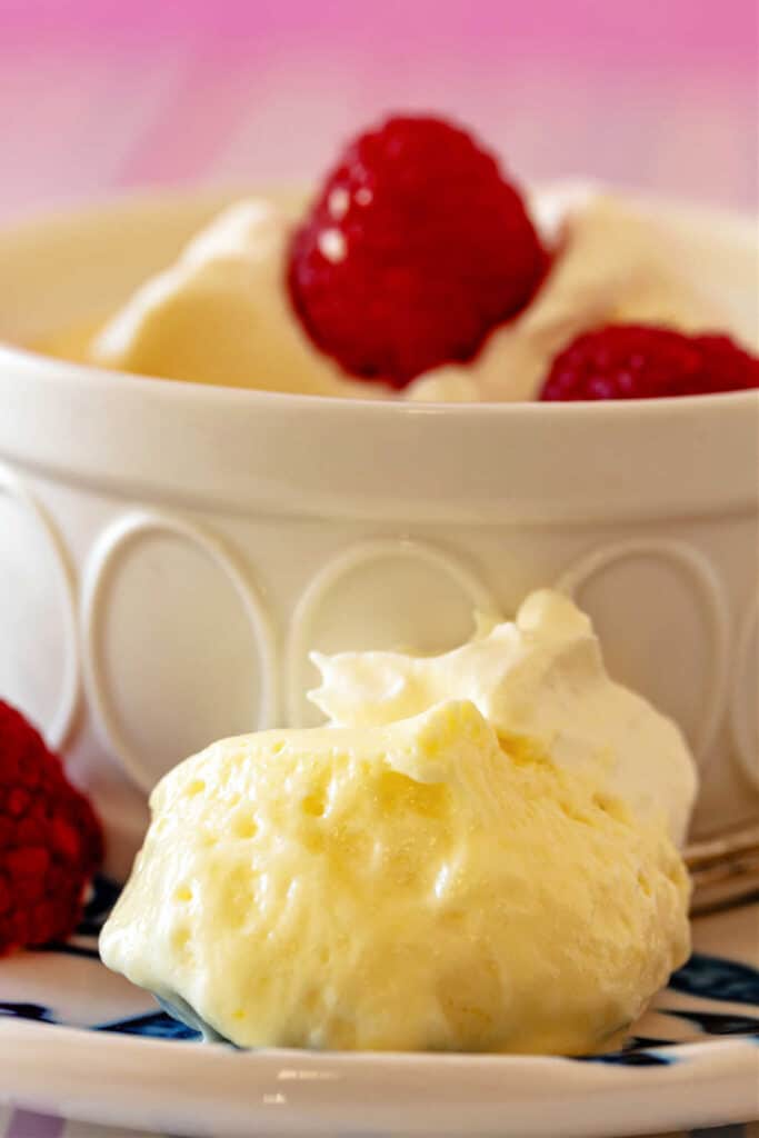 Close-up of a white ramekin of lemon mousse with a raspberry on top. In front of the ramekin is a bite of the mousse on a spoon showing the light and fluffy texture.
