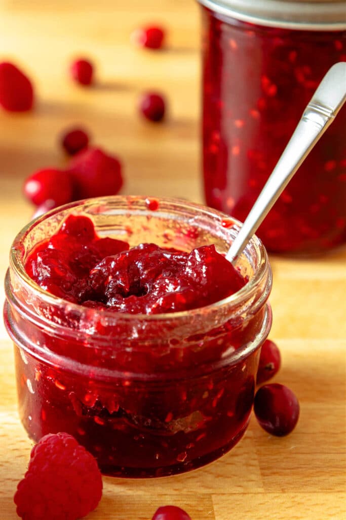 A small glass jar of red, cranberry raspberry jam with a small silver spoon in it. There is another lidded jar of jam in the background as well as a scattering of fresh cranberries and raspberries.