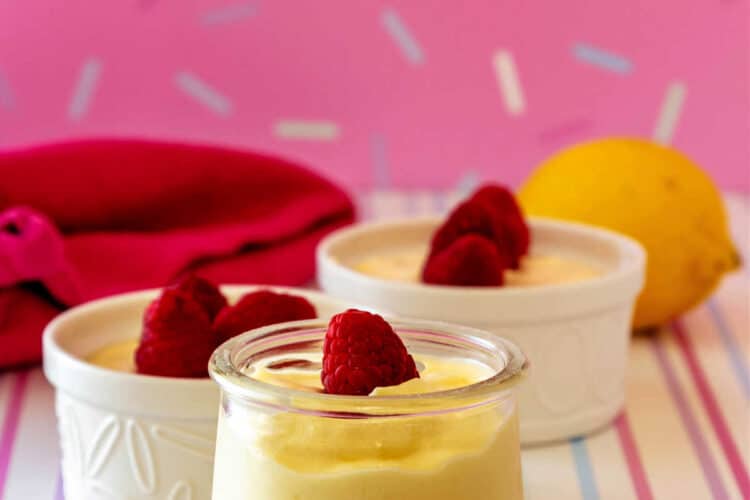 Two white ramekins and a small, glass jar of lemon mousse, each topped with a raspberry on a white striped surface with a pink background with sprinkles on it.