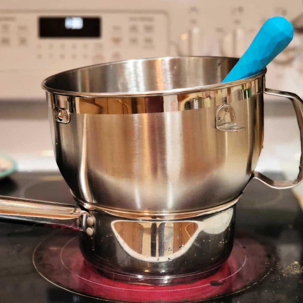 A metal mixing bowl set on top of a small saucepan. There's a blue-handled whisk sticking out of the bowl, and the burner is glowing red.