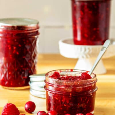 Three jars of red jam, one of which is open with a spoon in it. There are cranberries and a couple of raspberries scattered around on the counter near the open jar.