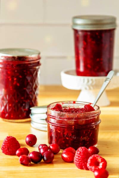Three jars of red jam, one of which is open with a spoon in it. There are cranberries and a couple of raspberries scattered around on the counter near the open jar.