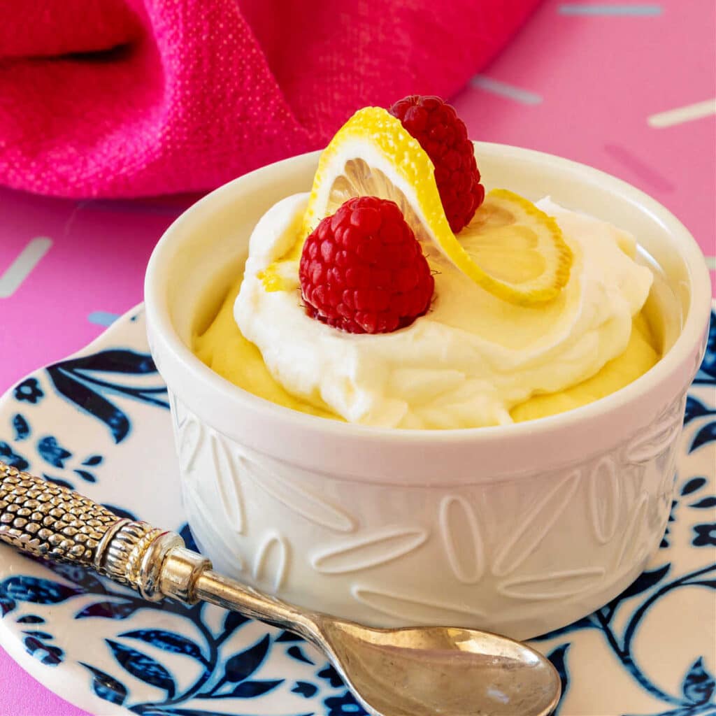 A close-up of a white ramekin of lemon mousse topped with whipped cream, a twist of lemon, and a couple of raspberries on a blue-and-whited patterned plate set on a bright pink surface.
